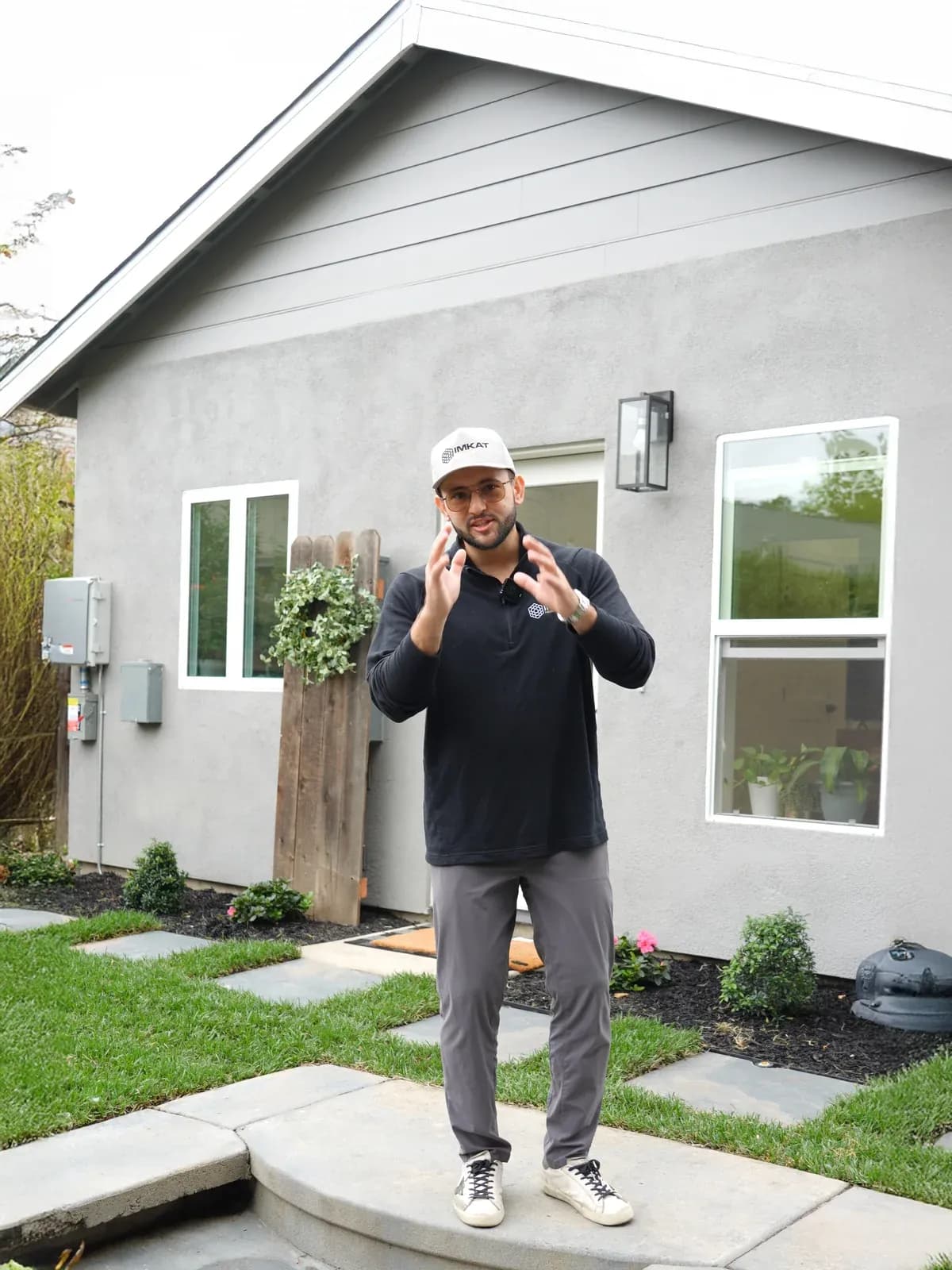 A man in an IMKAT cap talking to camera outside a house