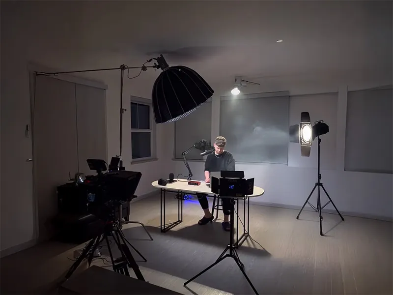 A man at a desk in a studio surrounded by lighting and camera equipment.